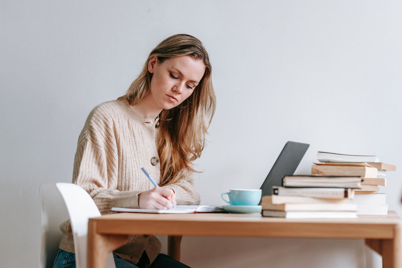 A woman writing in a notebook beside a laptop and a stack of books.