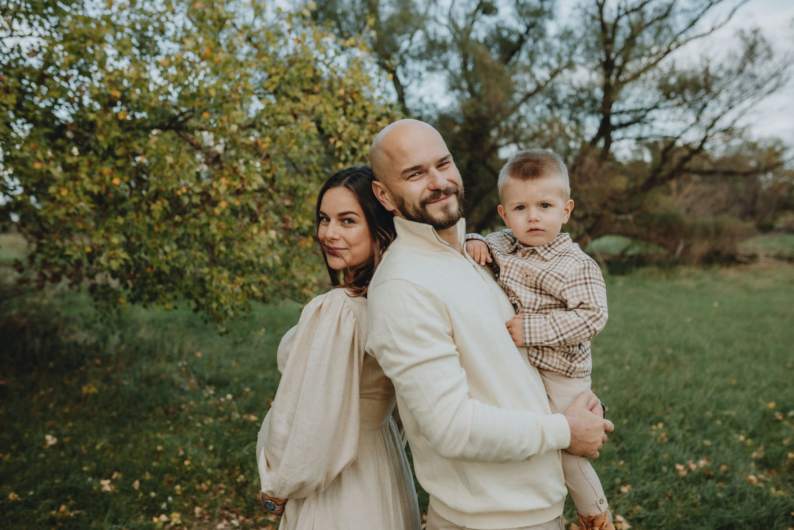 Parents and their young child posing outdoors beneath autumn trees.