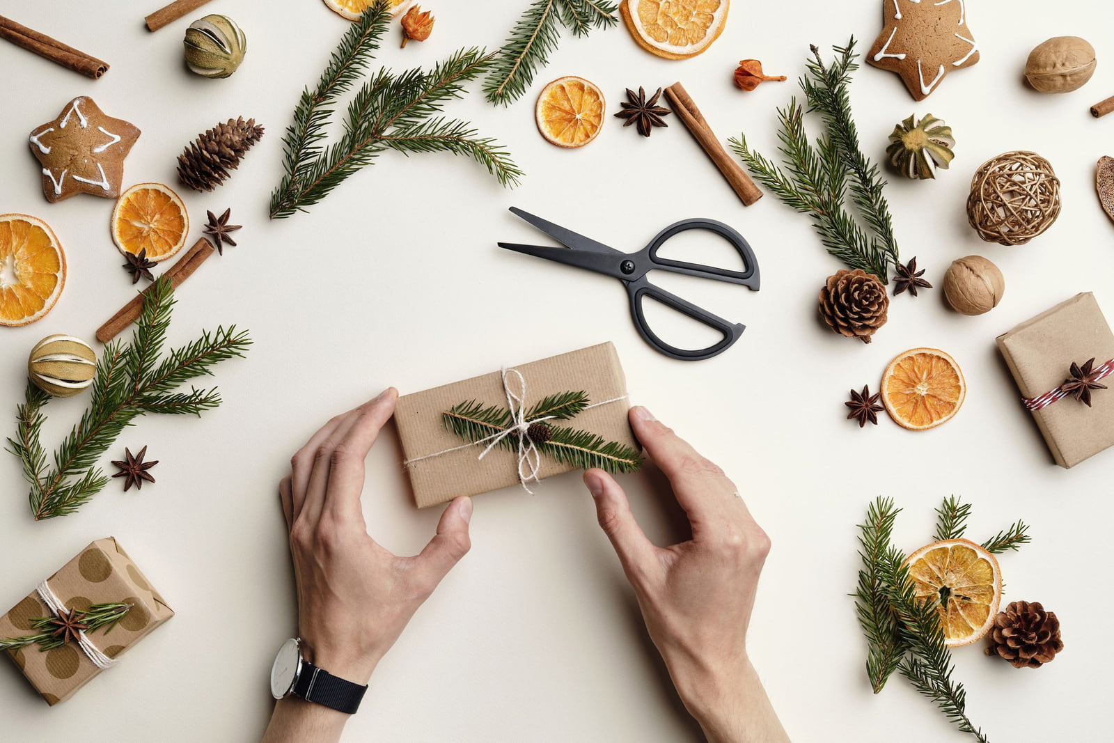 Hands tying a wrapped gift surrounded by evergreen sprigs, dried orange and festive decorations.