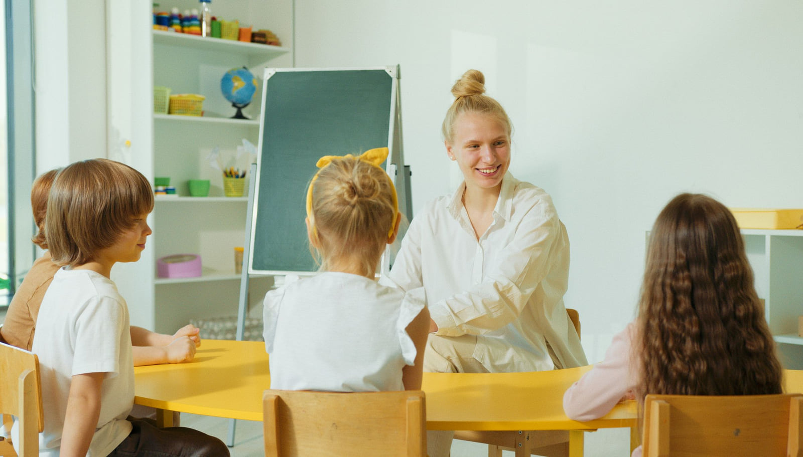 A preschool teacher sitting with young children around a low classroom table.