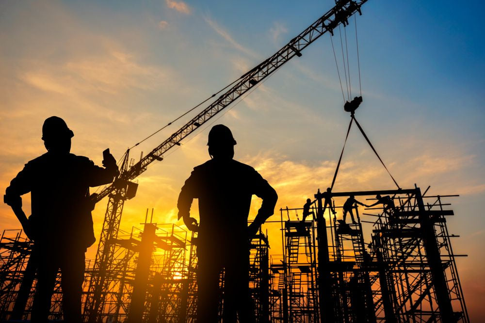 Two construction workers silhouetted against a sunset beside a tower crane.