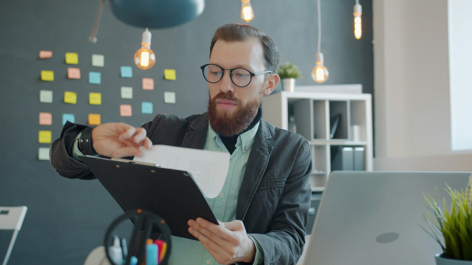 A professional checking papers on a clipboard beside a laptop.