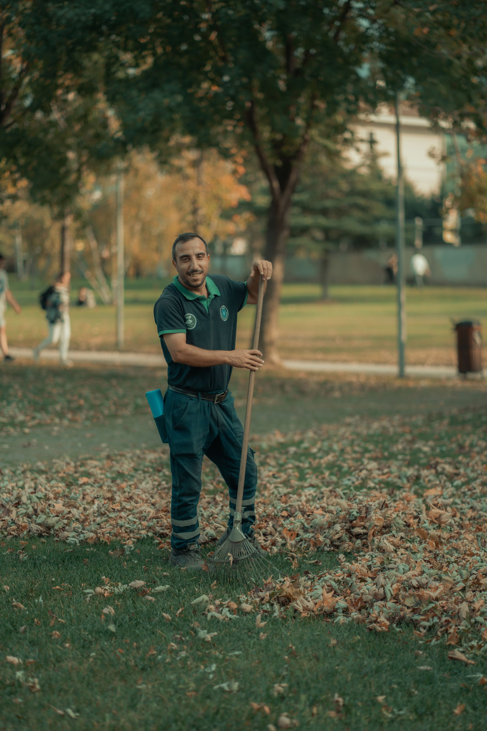 A worker holding rakes in a park covered with autumn leaves.