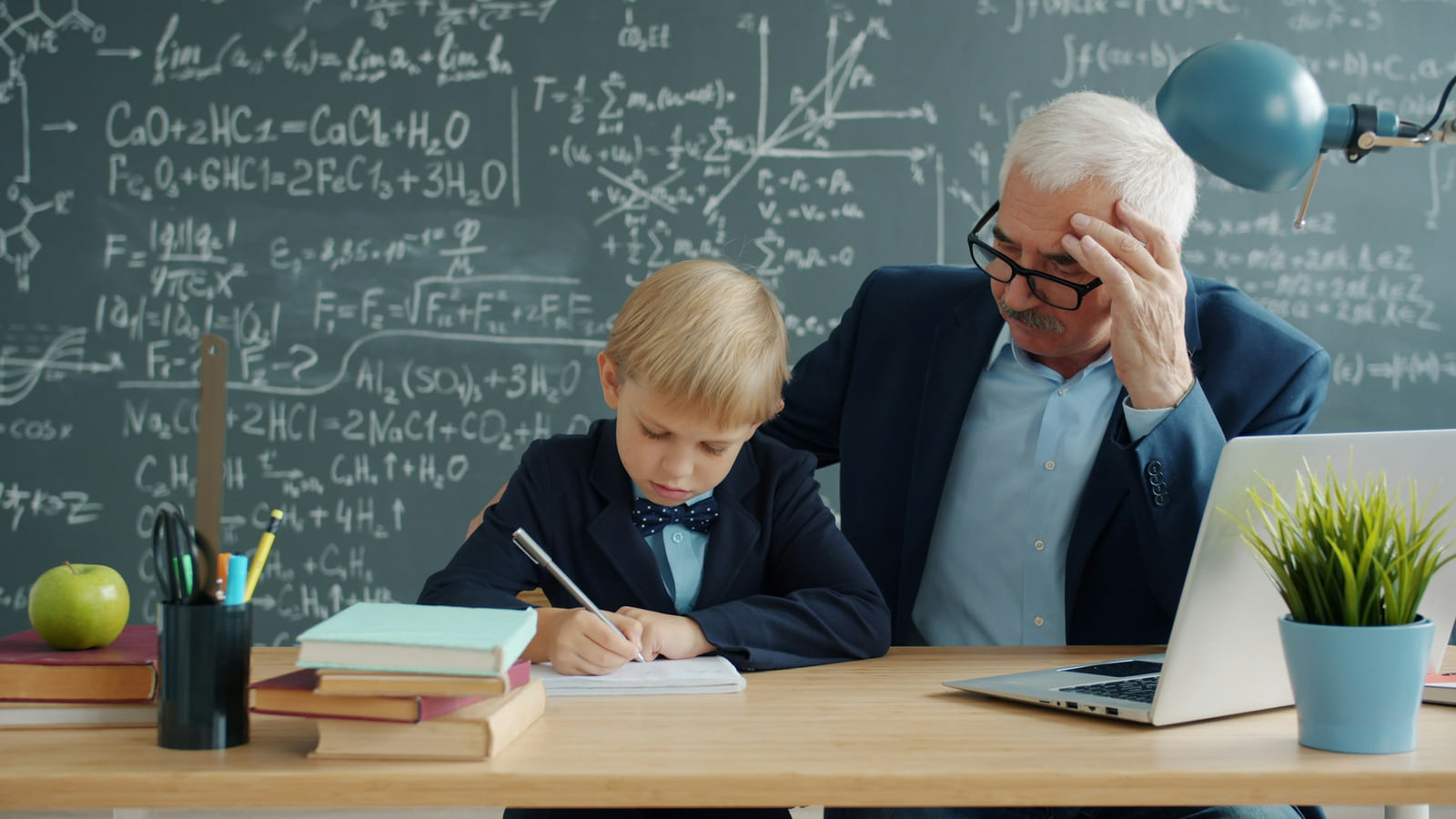 A teacher helping a young student write at a desk in front of a chalkboard.