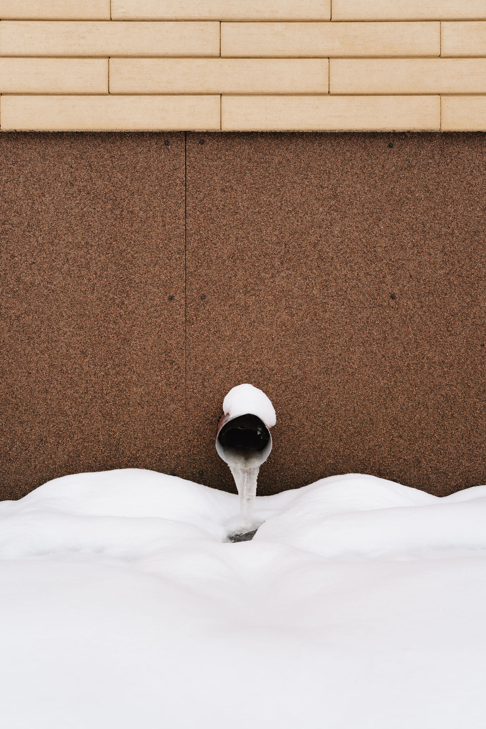 A wall pipe outlet above deep snow beside a house.