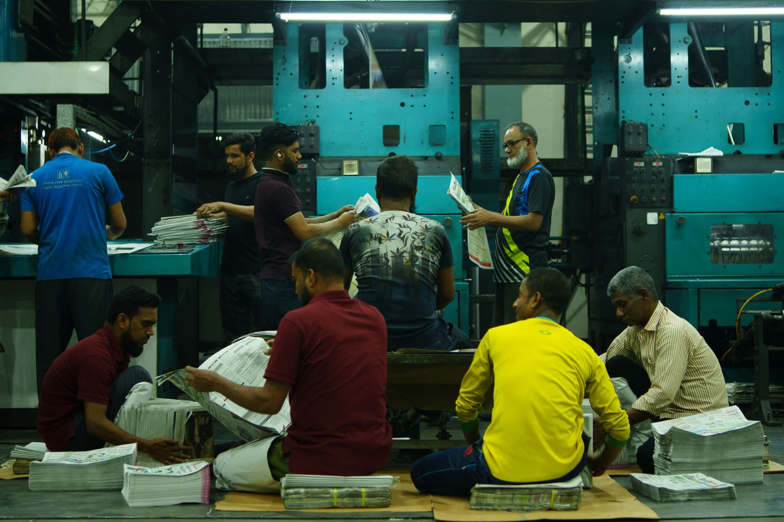 Workers handling printed sheets beside large printing equipment.