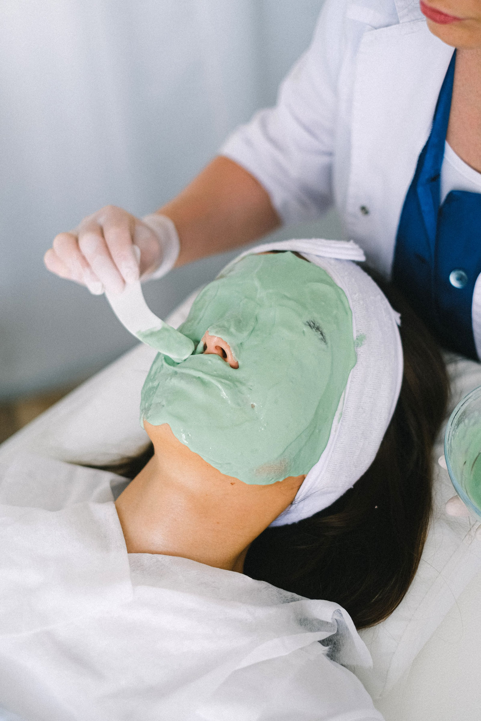An esthetician applying a green facial mask to a reclining client.