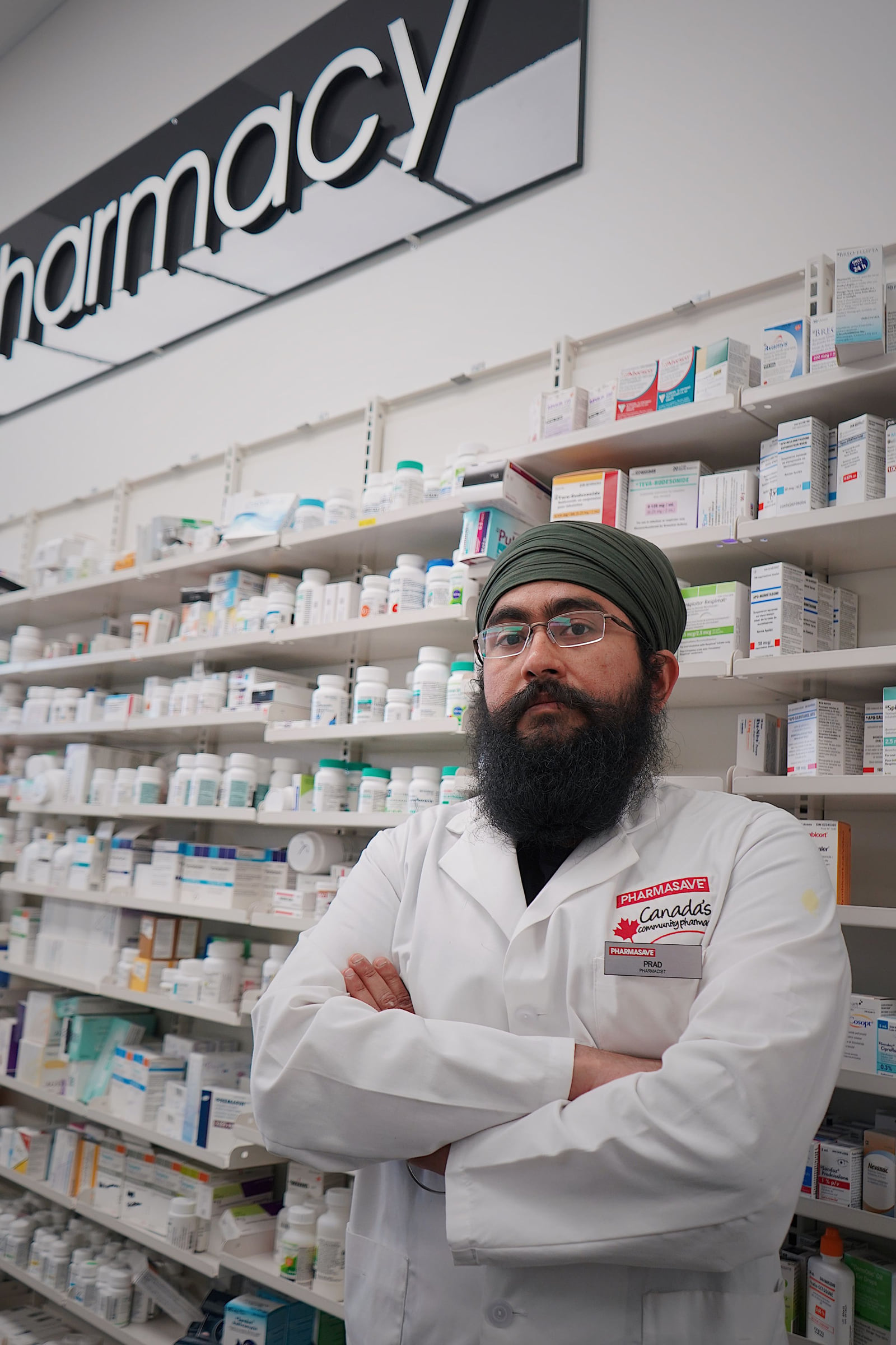 A pharmacist standing with folded arms in front of pharmacy shelves.