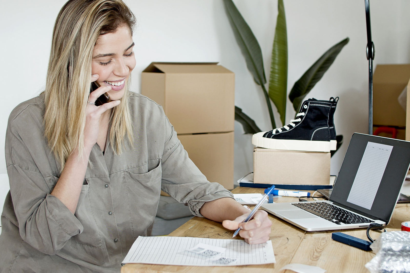A small-business owner taking a phone call and writing notes beside a laptop.