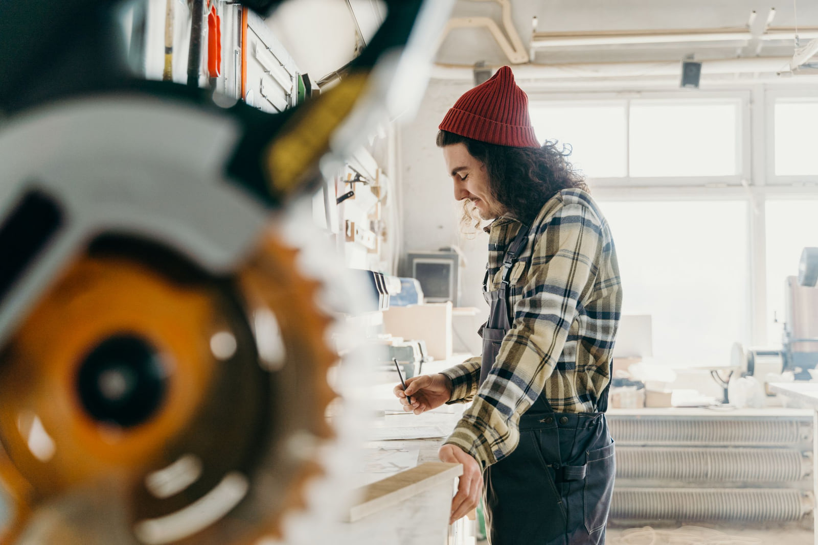 A carpenter measuring a board beside a saw in a bright workshop.