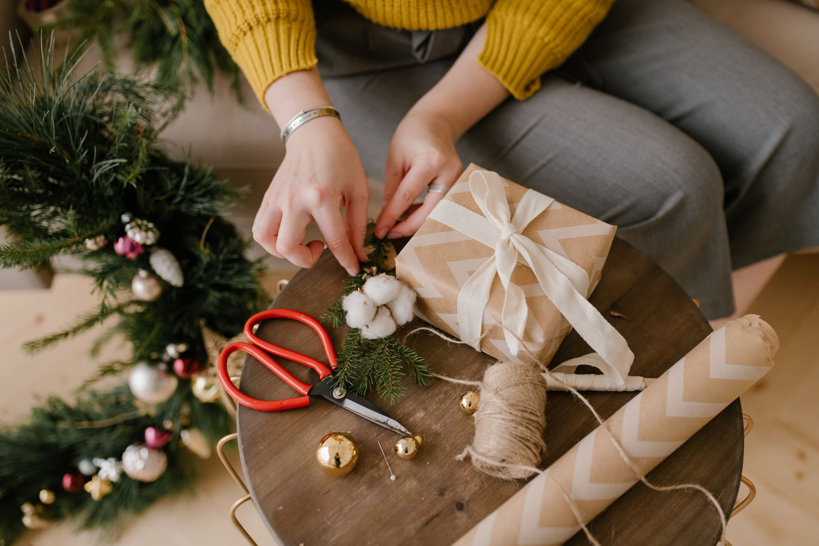 Hands wrapping a holiday gift beside evergreen branches and decorations.