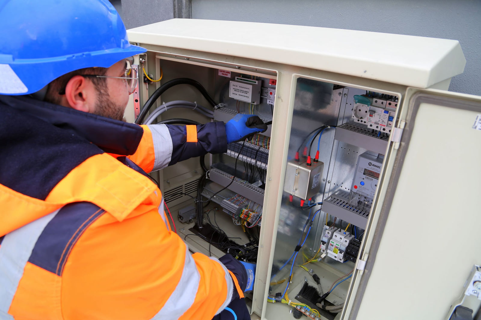 A worker in a hard hat and protective clothing inspecting an electrical cabinet.