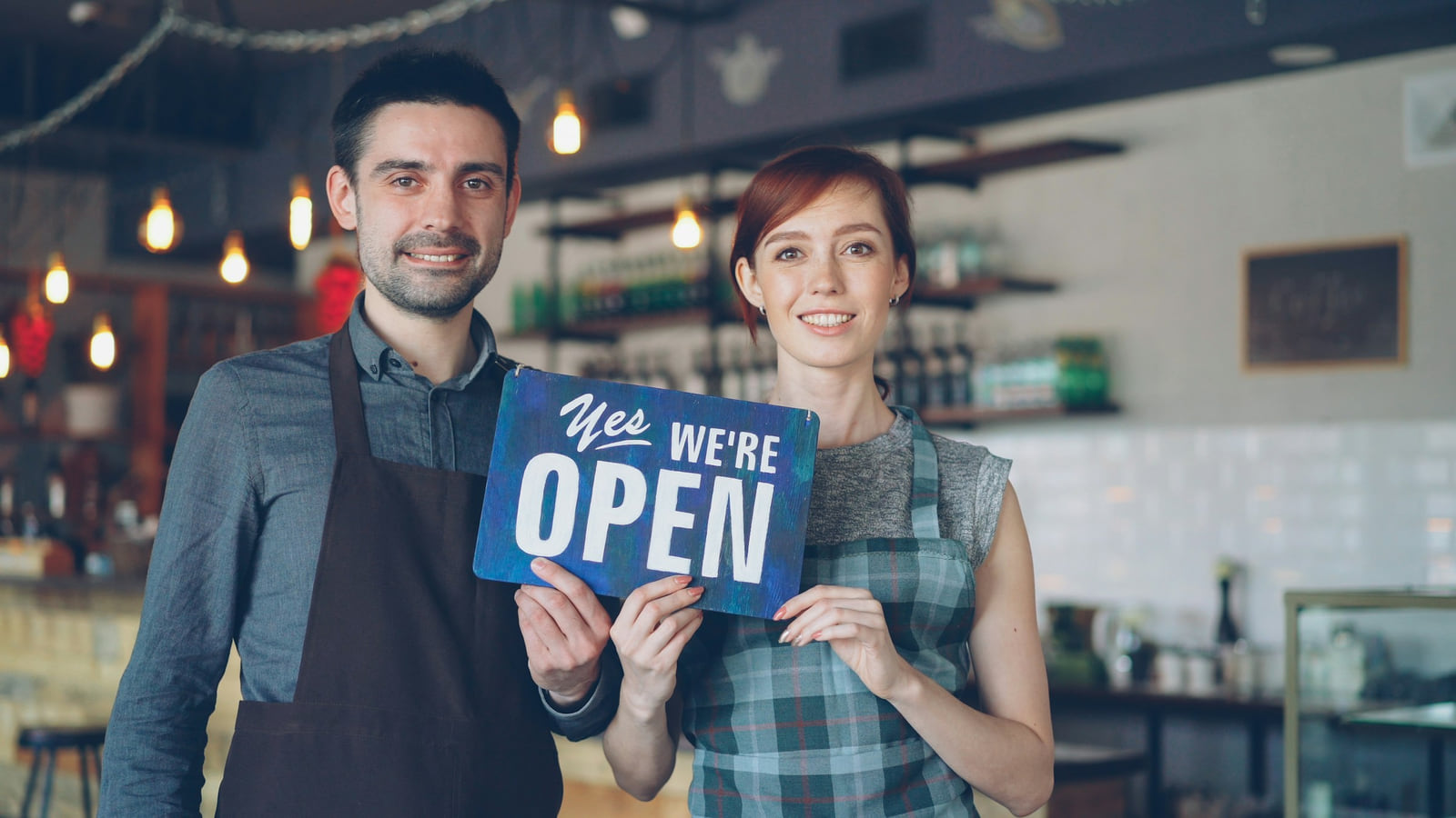 Two café owners holding an open sign inside their shop.