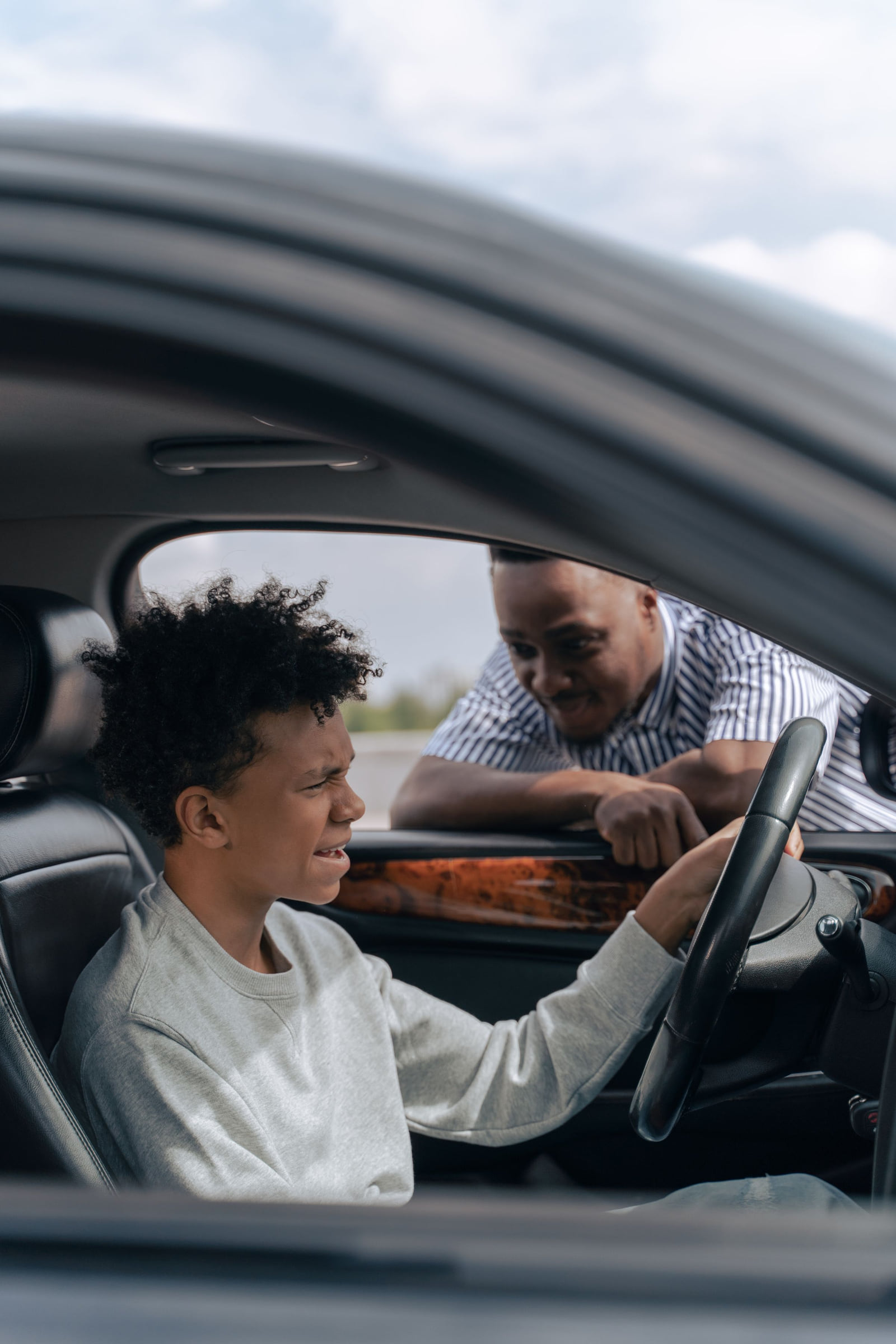 An adult guiding a teenager seated behind a car's steering wheel.
