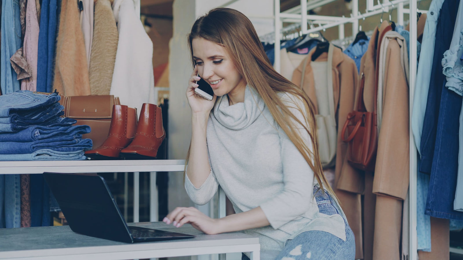 A woman using a laptop and phone behind the counter of a clothing shop.