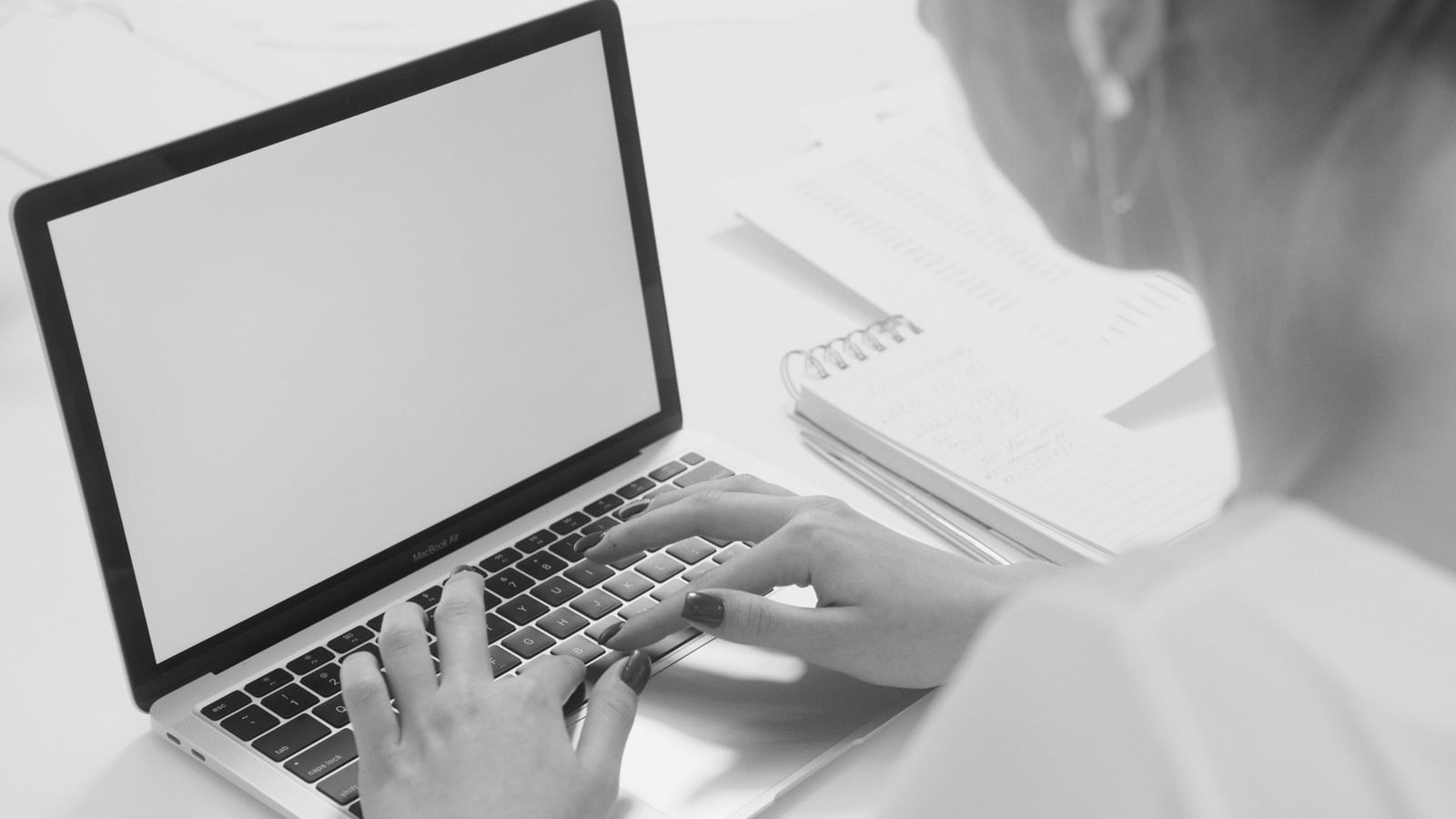 A person typing on a laptop beside an open notebook.