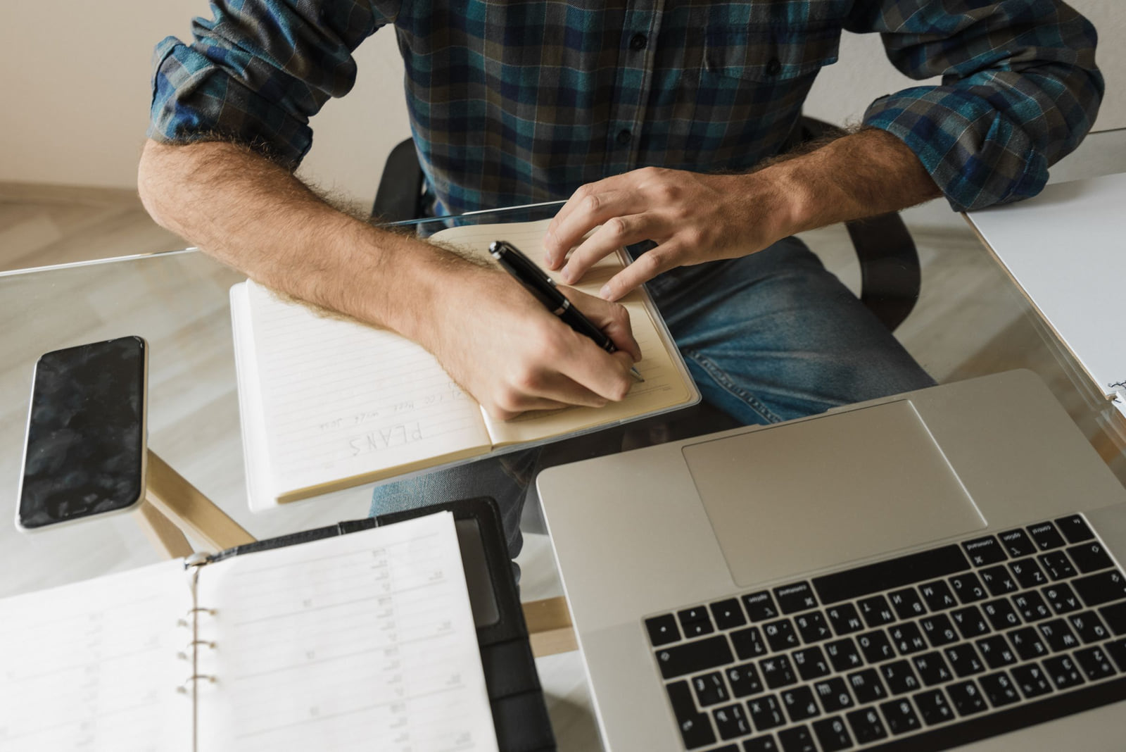 A person writing in a notebook beside an open laptop and phone.