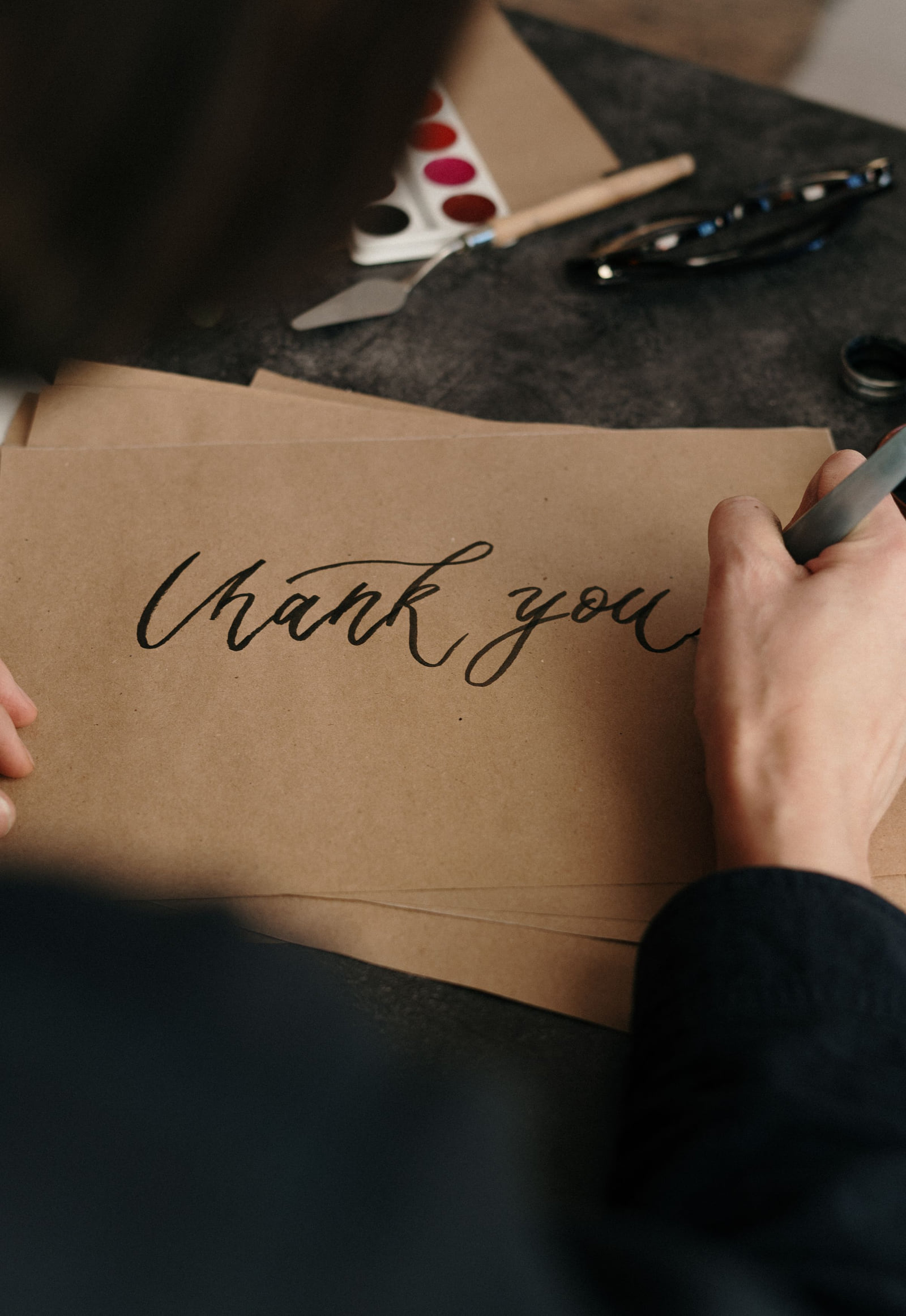 A person hand-lettering Thank You on a sheet of brown paper.