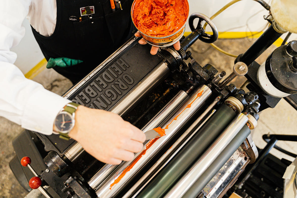 Hands operating a traditional printing press with rollers and metal type.
