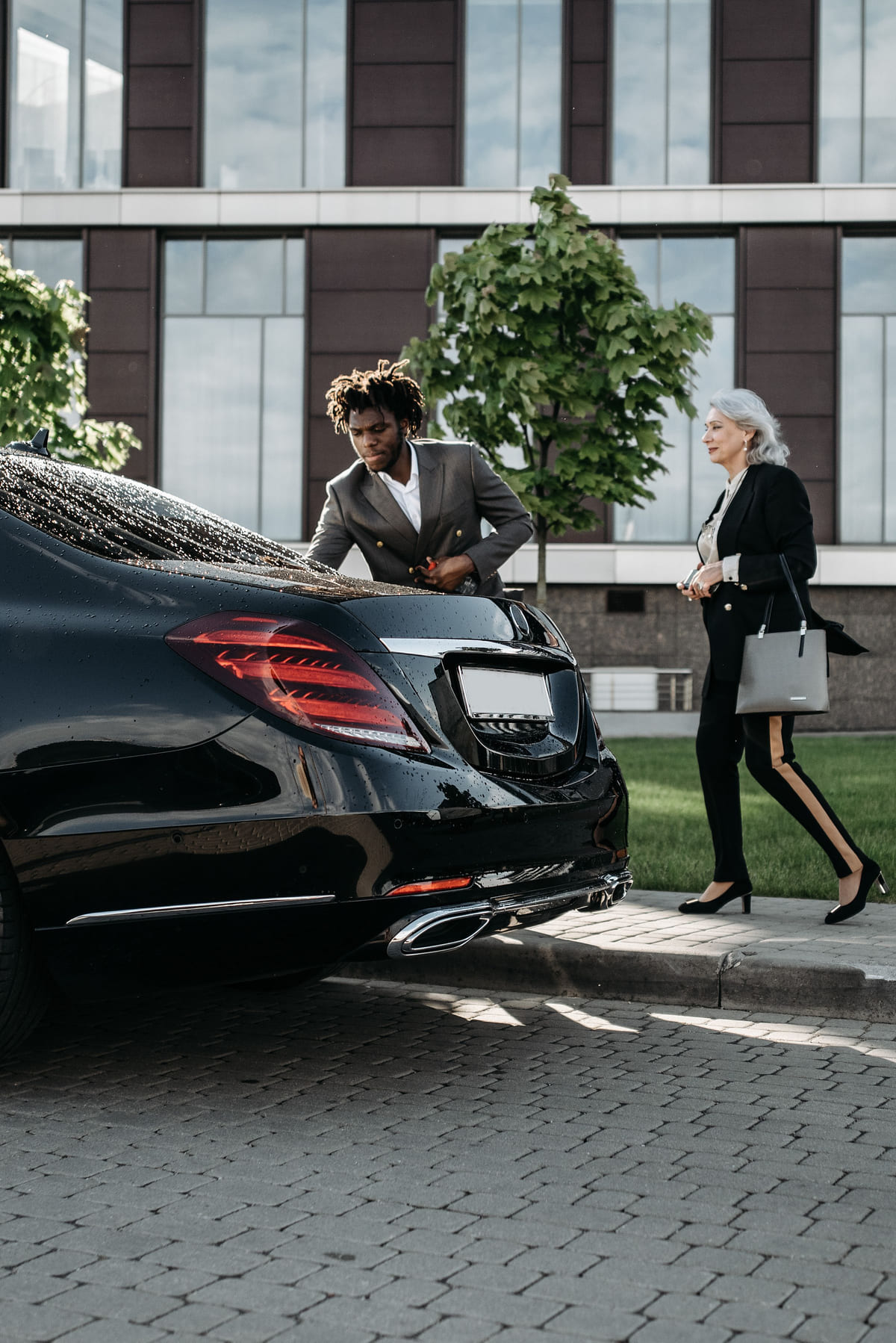 A suited driver beside a black sedan as a passenger approaches.