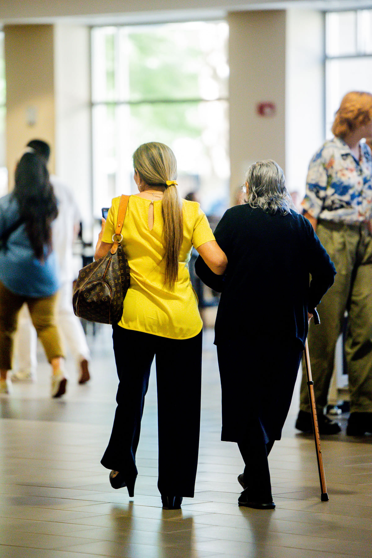 A caregiver supporting an older woman as they walk through a public space.