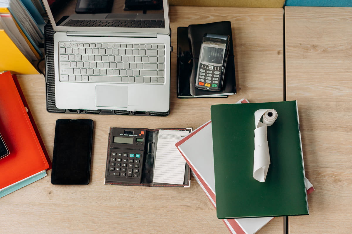 A laptop, calculator, payment terminal and financial paperwork arranged on a desk.