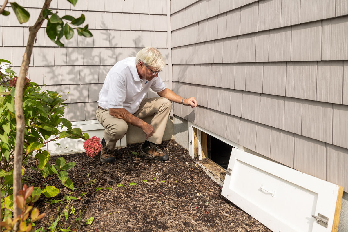 A home inspector checking a crawl-space opening beside a house.