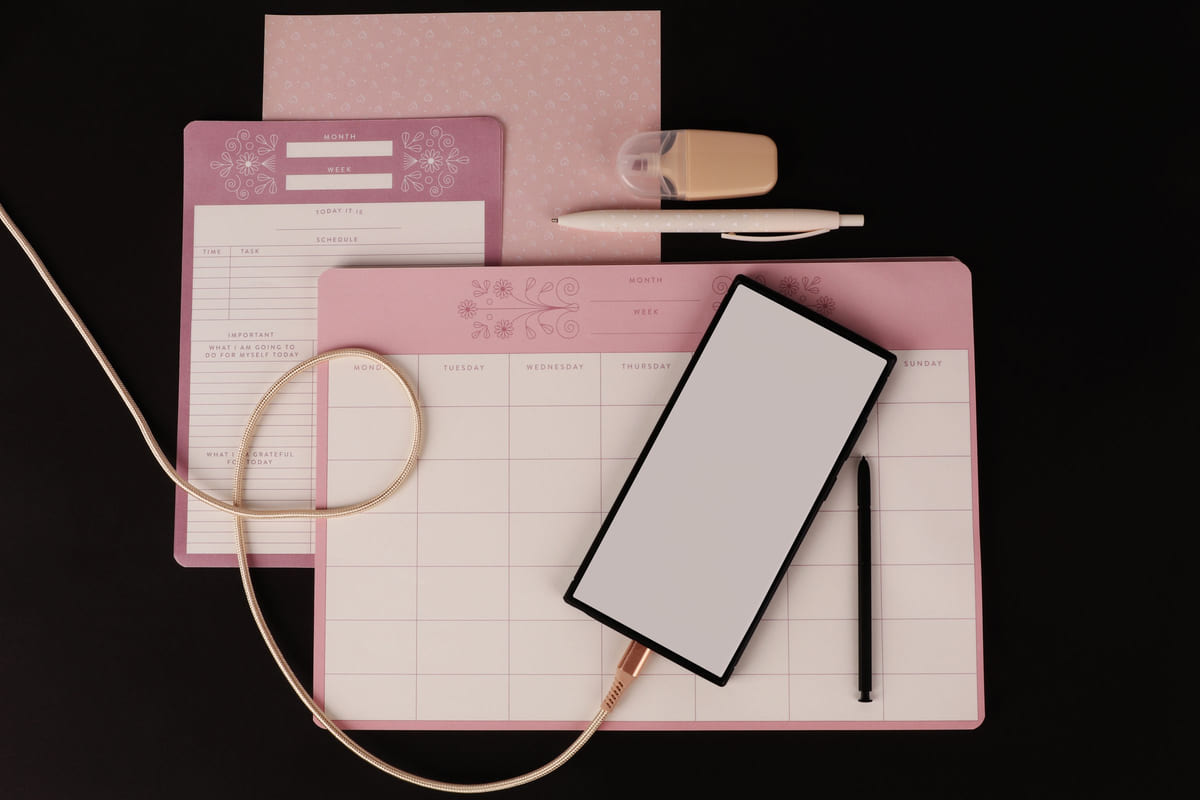 A tablet, paper planner, pen and stationery arranged on a pink desk.