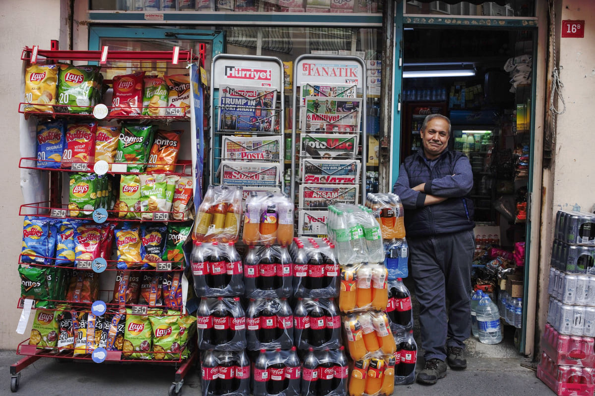 A shopkeeper beside a small grocery entrance surrounded by displayed goods.