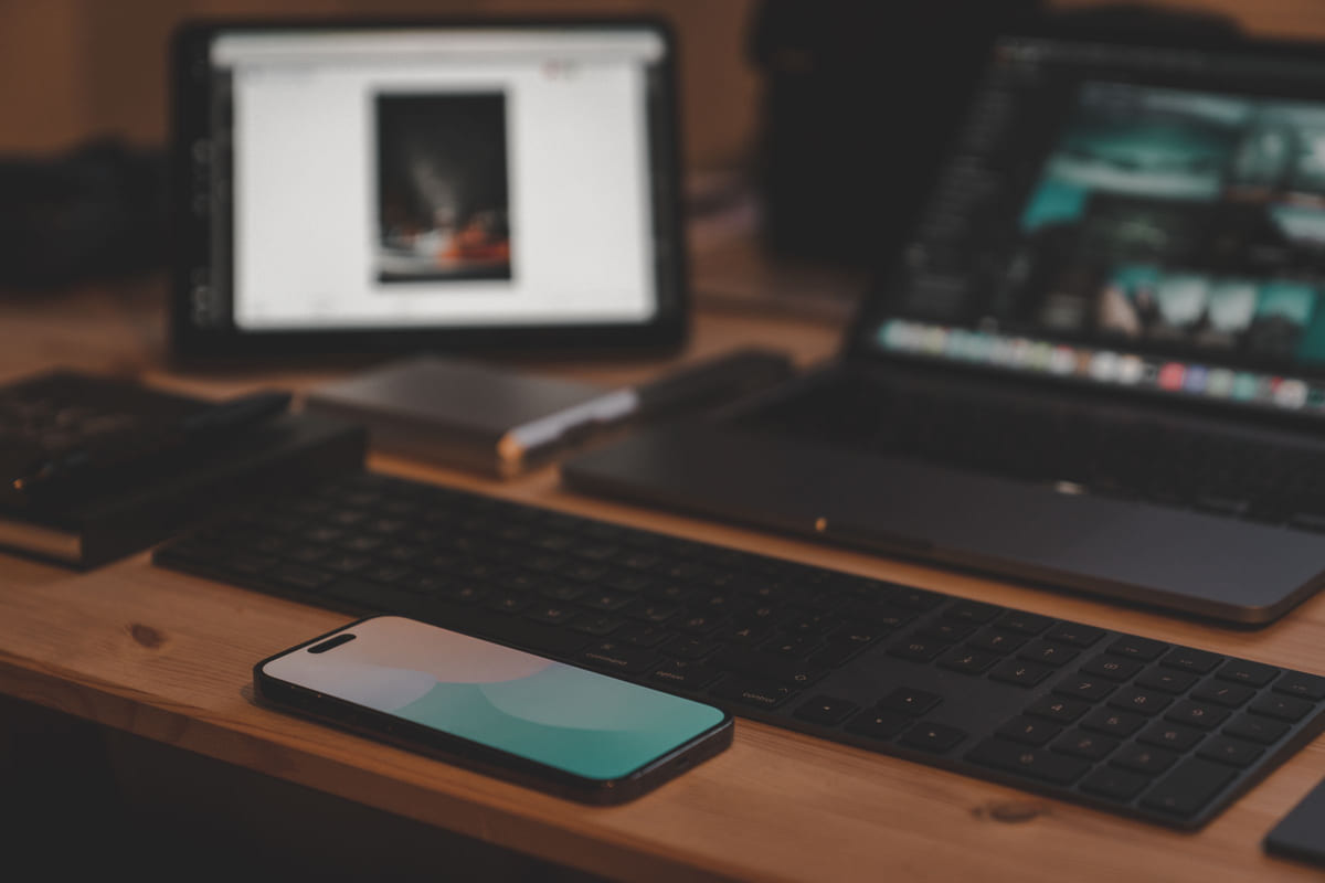 A smartphone, keyboard, laptop and tablet arranged on a wooden desk.