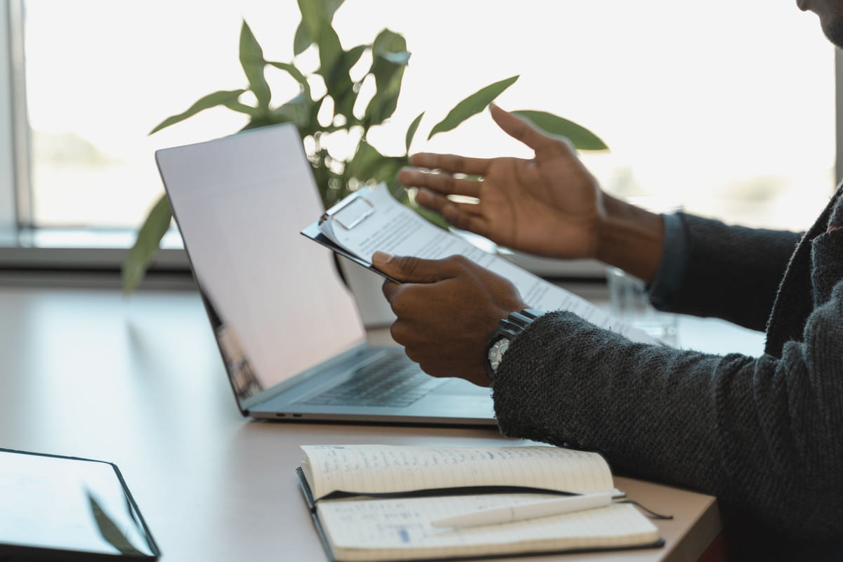 A person checking documents on a clipboard beside a laptop and notebook.