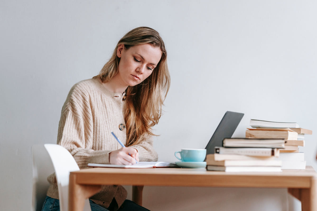 A woman writing in a notebook beside a laptop and a stack of books.