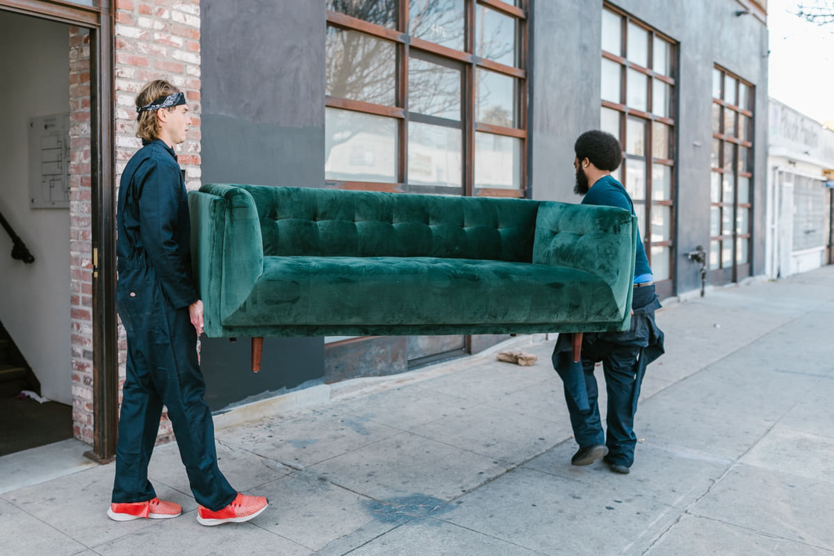 Two workers carrying a large green sofa outside a building.