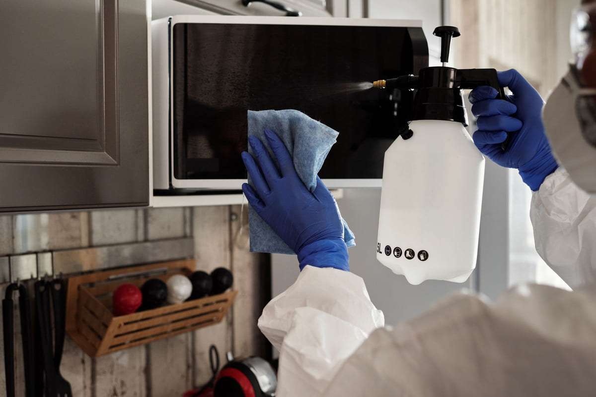 Gloved hands wiping the door of a microwave in a kitchen.