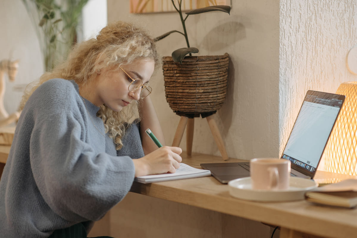 A woman writing in a notebook beside an open laptop and a mug.