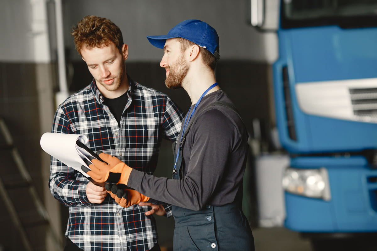 A mechanic and customer reviewing paperwork beside a car in a workshop.