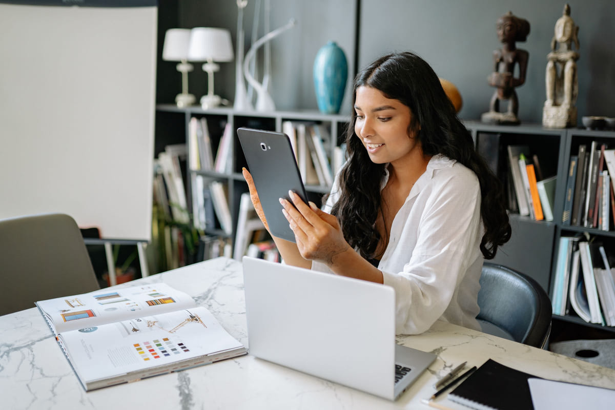 A woman using a tablet beside a laptop and printed design materials.