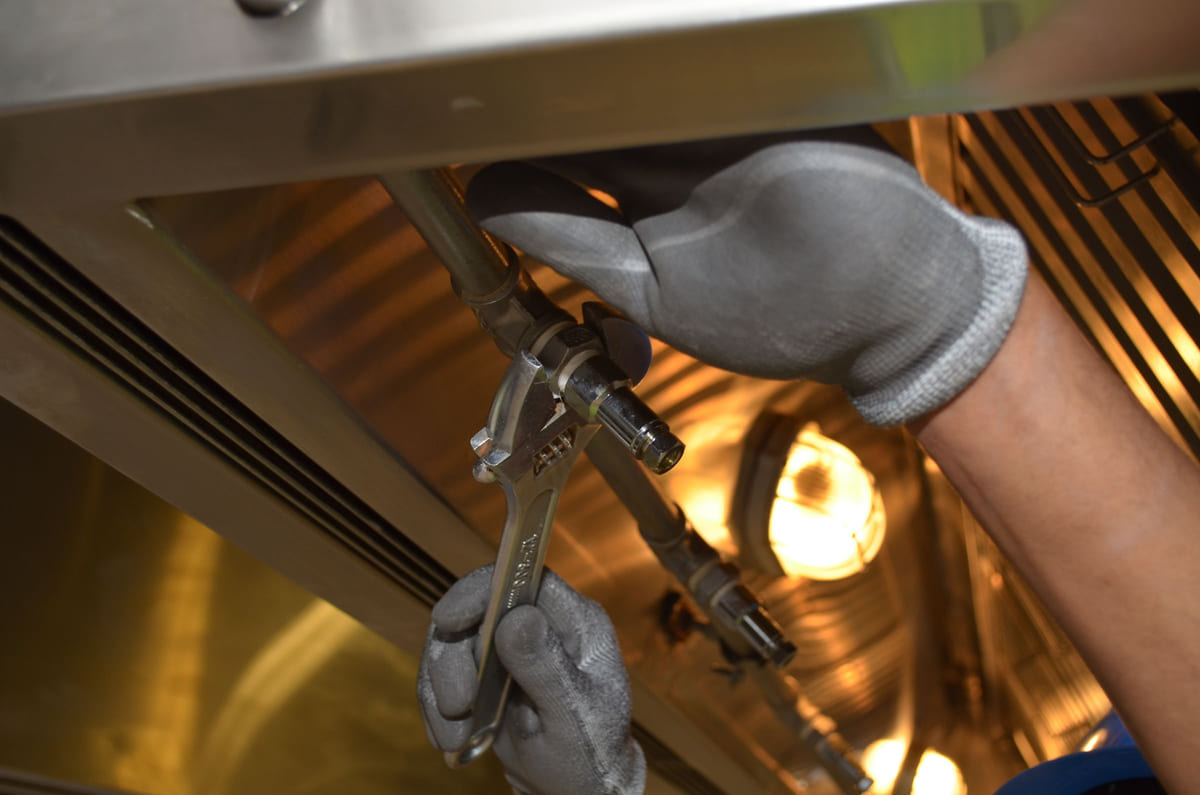 Gloved hands using a wrench on overhead pipework.