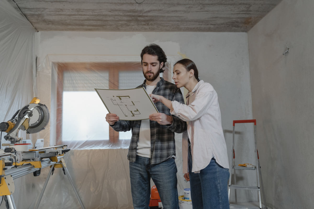 Two people reviewing a floor plan in a room undergoing renovation.