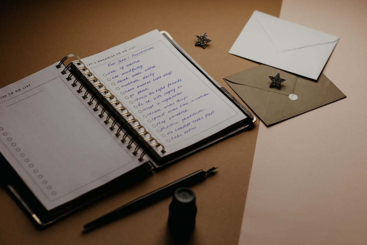 An open planner, handwritten checklist, envelopes and a pen on a desk.