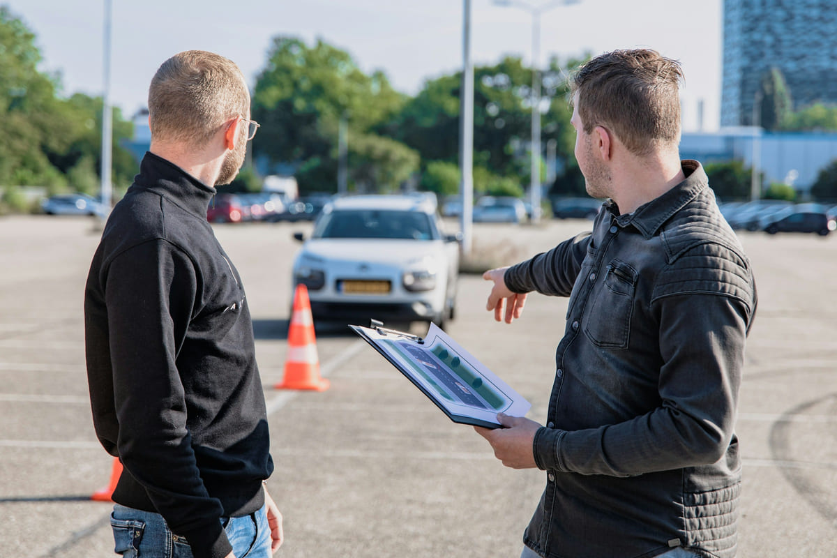 A driving instructor using a clipboard while explaining a practice area beside a car.