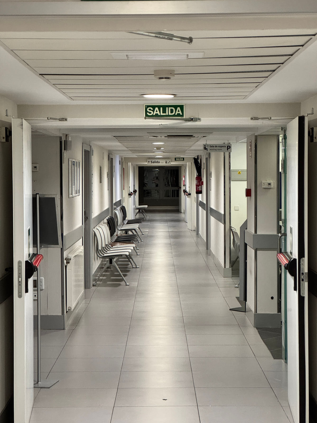 An empty hospital corridor with doors, handrails and an exit sign.