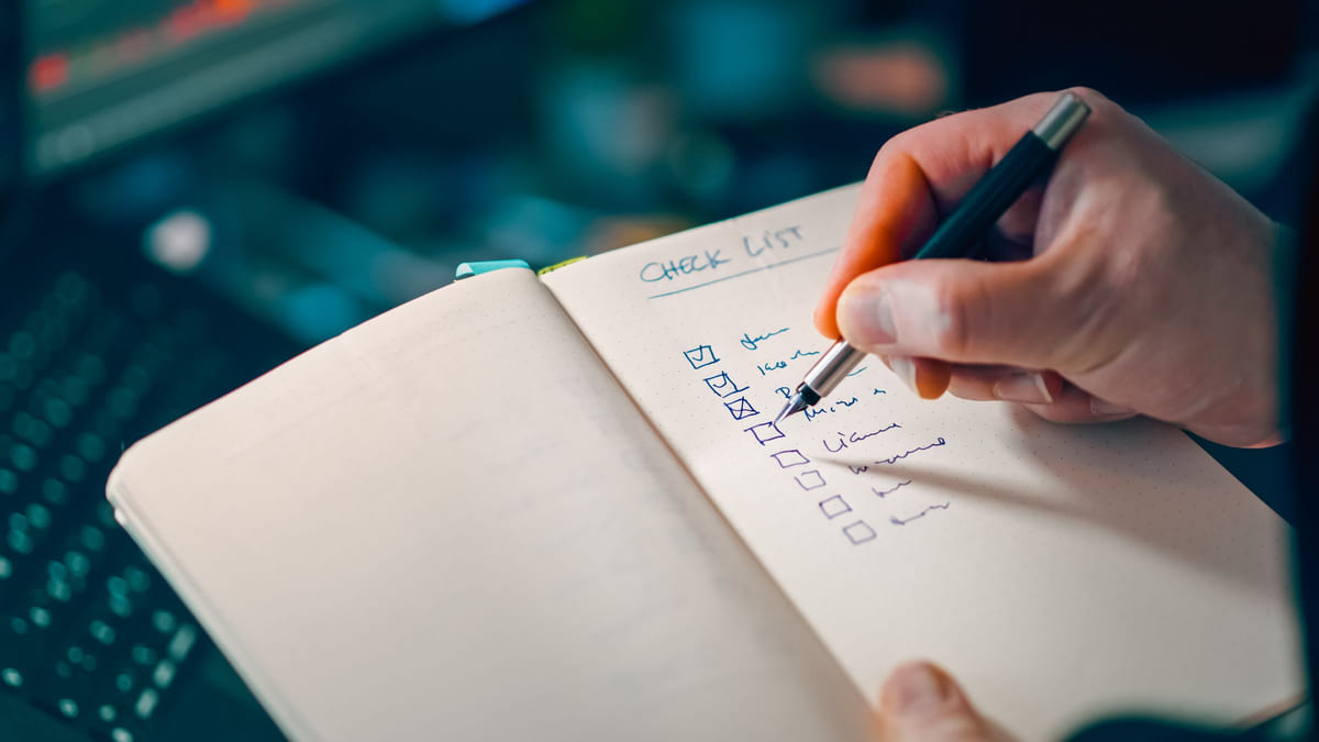 A hand ticking items in a handwritten checklist beside a laptop.