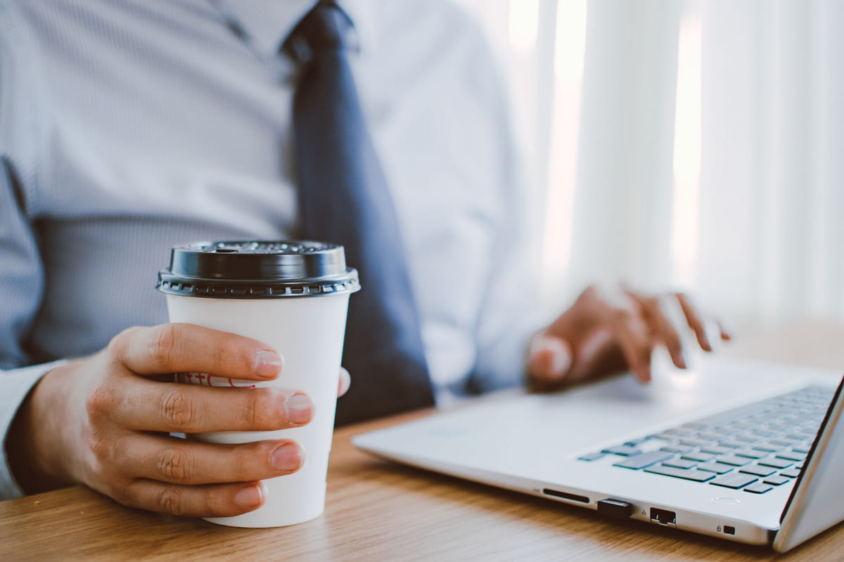 A person holding a takeaway coffee beside an open laptop.