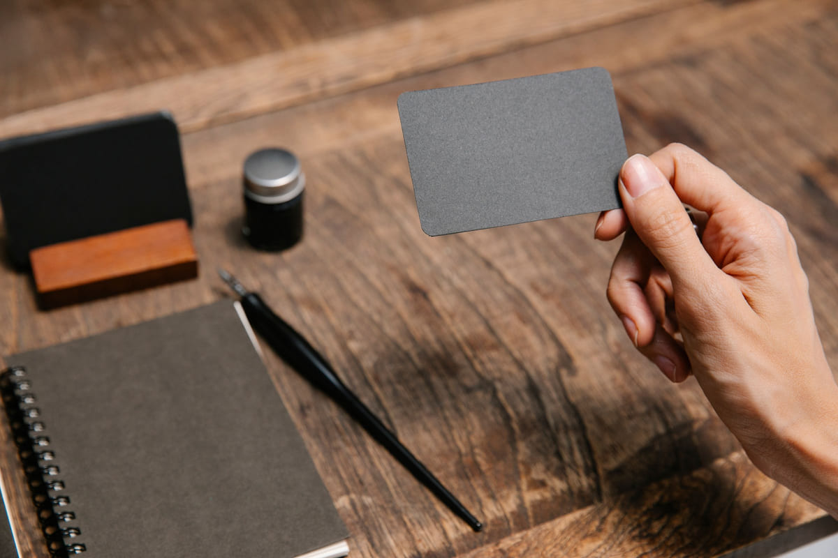 A hand holding a plain dark business card above a wooden desk.