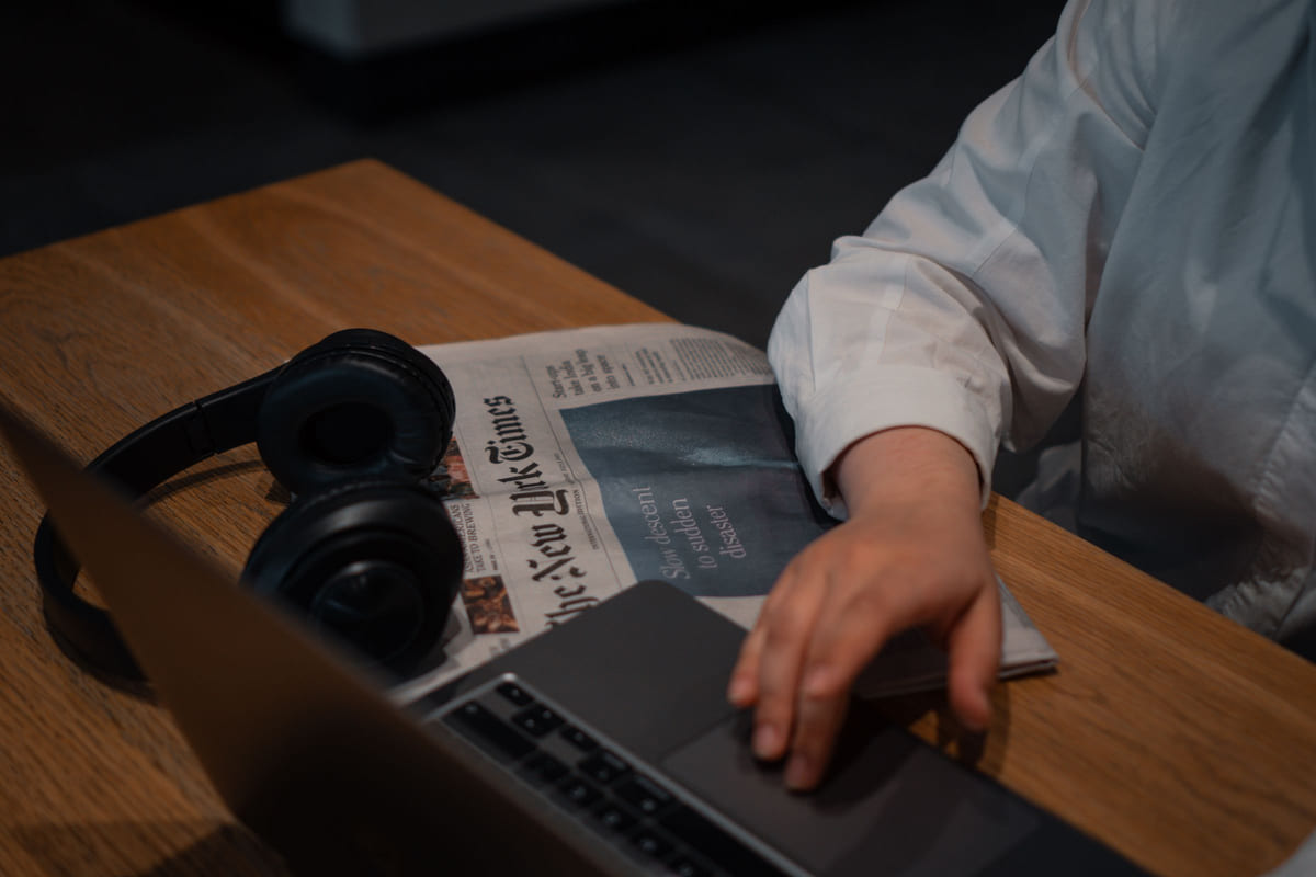 A person using a laptop beside a newspaper and headphones.